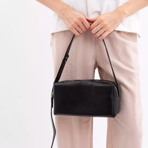 Pu&ograve; includere: Una borsa a tracolla rettangolare in pelle nera con finitura liscia, dotata di chiusura superiore con zip e una tracolla sottile regolabile. La borsa presenta una silhouette strutturata e squadrata con linee pulite.