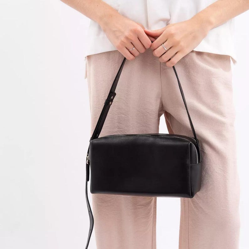 Pu&ograve; includere: Una borsa a tracolla rettangolare in pelle nera con finitura liscia, dotata di chiusura superiore con zip e una tracolla sottile regolabile. La borsa presenta una silhouette strutturata e squadrata con linee pulite.