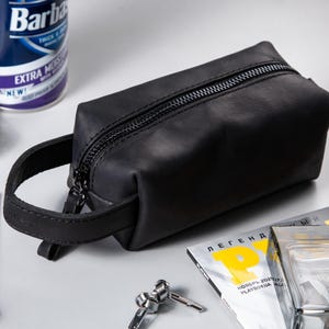 May include: A rectangular black leather wash bag featuring a top zip closure and a side carry handle. The bag has a smooth, matte finish and is constructed with visible stitching along the edges and the handle attachment point.