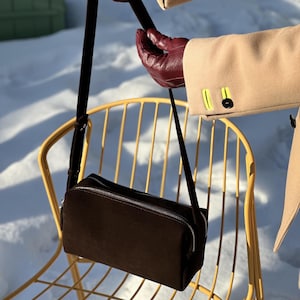 Pu&ograve; includere: Una borsa a tracolla rettangolare in camoscio marrone scuro con chiusura a zip in metallo argentato e tracolla regolabile in tinta. La borsa presenta un design strutturato e pulito con una texture liscia e un'estetica minimalista.