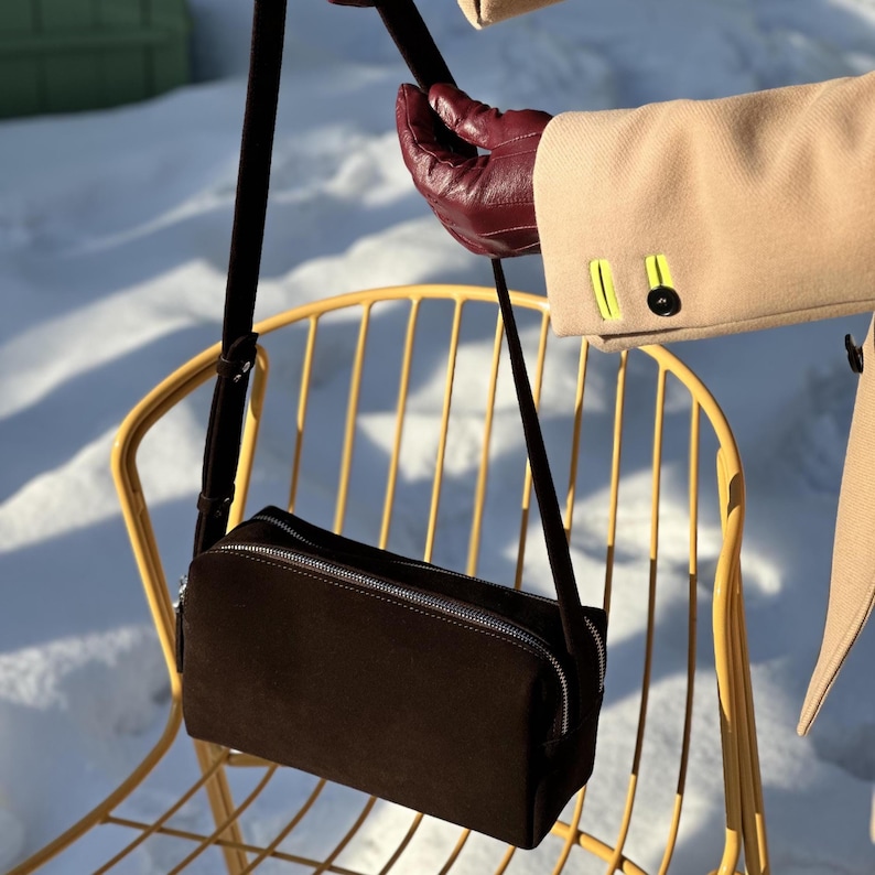 Pu&ograve; includere: Una borsa a tracolla rettangolare in camoscio marrone scuro con chiusura a zip in metallo argentato e tracolla regolabile in tinta. La borsa presenta un design strutturato e pulito con una texture liscia e un'estetica minimalista.