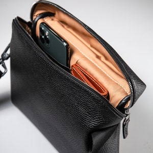 May include: A black pebbled leather crossbody bag with a zippered top opening. The interior is lined with tan fabric and contains a dark-colored smartphone and a small brown leather card holder tucked into the main compartment.