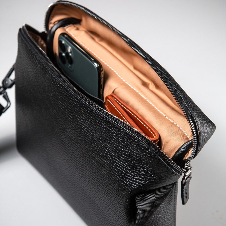 May include: A black pebbled leather crossbody bag with a zippered top opening. The interior is lined with tan fabric and contains a dark-colored smartphone and a small brown leather card holder tucked into the main compartment.