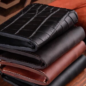 May include: A stack of four leather wallets. The top wallet is black with a textured crocodile skin pattern. The three wallets underneath are smooth, featuring shades of dark brown, medium brown, and black, with visible stitching along the edges.