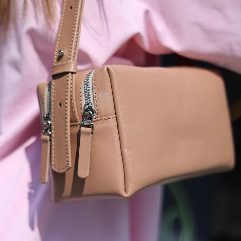 May include: A rectangular tan leather shoulder bag featuring a smooth finish, a wide adjustable strap with a silver metal stud, and two silver-toned metal zips with matching leather pull tabs on the side of the main compartment.