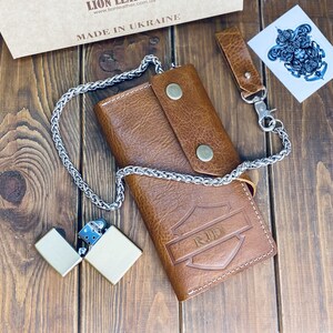 Puede incluir: Una cartera de cuero color canela con un dise&ntilde;o en relieve en forma de escudo con las letras RJD, asegurada por dos botones a presi&oacute;n color lat&oacute;n. Una cadena de metal plateado est&aacute; unida a la cartera, junto a un encendedor de metal a juego.