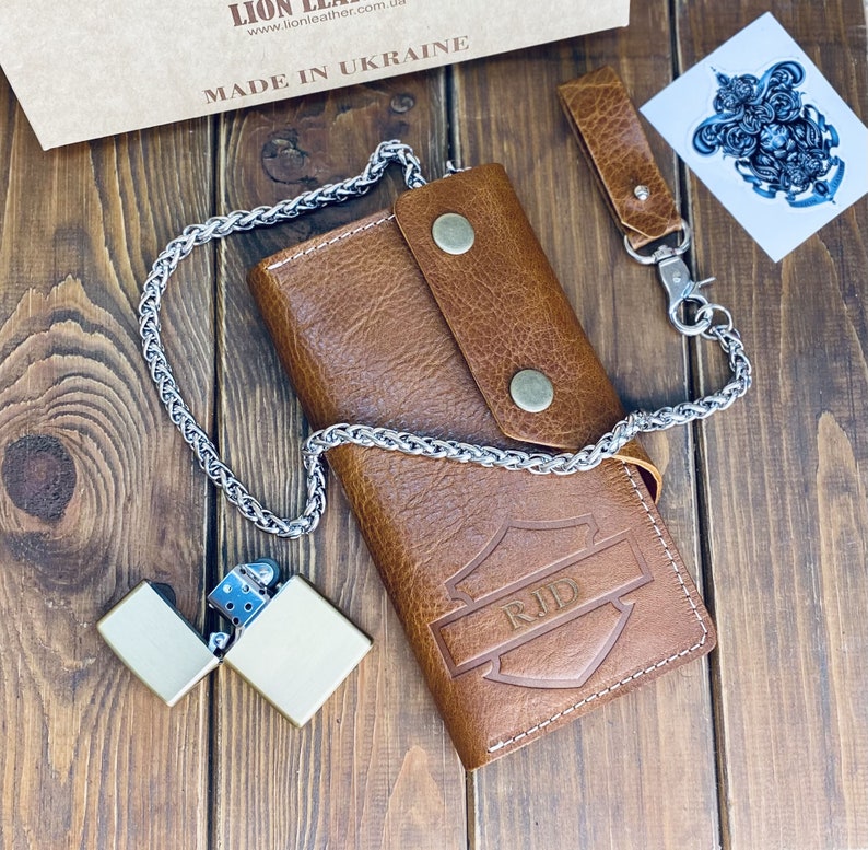 Puede incluir: Una cartera de cuero color canela con un dise&ntilde;o en relieve en forma de escudo con las letras RJD, asegurada por dos botones a presi&oacute;n color lat&oacute;n. Una cadena de metal plateado est&aacute; unida a la cartera, junto a un encendedor de metal a juego.