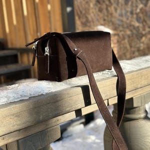 Pu&ograve; includere: Una borsa a tracolla rettangolare in camoscio marrone scuro con una consistenza liscia, una lunga tracolla regolabile e cerniere in metallo color argento con tiretti in camoscio marrone abbinati. La borsa ha una forma strutturata e squadrata.