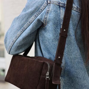 Pu&ograve; includere: Una borsa a tracolla rettangolare in camoscio marrone scuro con chiusura a zip in metallo argentato e tracolla regolabile abbinata con dettagli in argento, indossata sopra una giacca di jeans azzurro slavato con cuciture visibili.