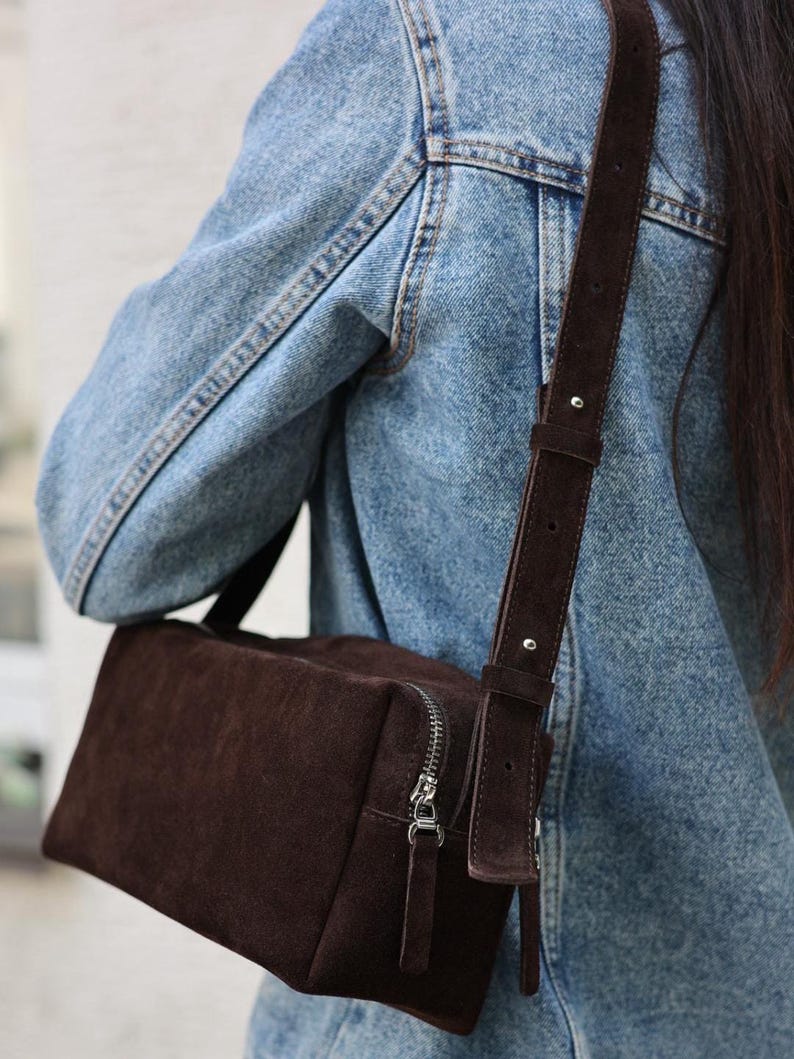 Pu&ograve; includere: Una borsa a tracolla rettangolare in camoscio marrone scuro con chiusura a zip in metallo argentato e tracolla regolabile abbinata con dettagli in argento, indossata sopra una giacca di jeans azzurro slavato con cuciture visibili.