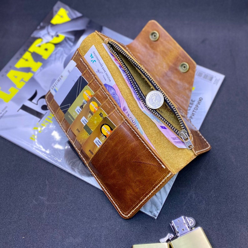 Puede incluir: Una cartera de cuero color canela con m&uacute;ltiples ranuras para tarjetas, un compartimento para monedas con cremallera y tirador plateado, y cierre de bot&oacute;n a presi&oacute;n. El interior contiene varias tarjetas y billetes con texto en ucraniano.