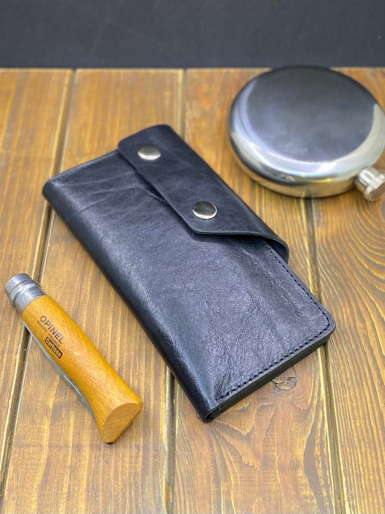 Puede incluir: Una cartera de cuero negro con una solapa plegable asegurada por dos broches de metal de color plateado. Junto a la cartera hay una navaja plegable con mango de madera clara marcada con el texto Opinel y una petaca redonda de metal plateado.