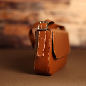 May include: A smooth, tan-colored leather shoulder bag featuring a structured rectangular body with rounded edges, a flap closure, and a silver-toned metal buckle on the adjustable strap, accented with visible light-colored stitching throughout.
