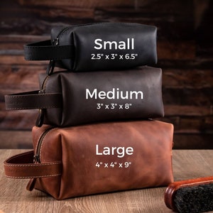 May include: Three rectangular leather toiletry bags stacked vertically in black, dark brown, and tan. Each bag features a side handle and zipper. Text labels indicate sizes: Small 6.35 cm x 7.62 cm x 16.51 cm, Medium 7.62 cm x 7.62 cm x 20.32 cm, and Large 10.16 cm x 10.16 cm x 22.86 cm.