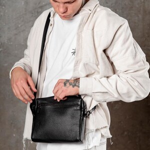 May include: A black pebbled leather crossbody bag with a rectangular shape, a top zipper closure, and a thin adjustable black shoulder strap. The bag is worn over a white t-shirt and a light beige corduroy button-down shirt with frayed hem details.