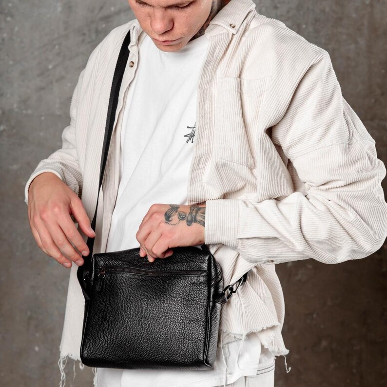 May include: A black pebbled leather crossbody bag with a rectangular shape, a top zipper closure, and a thin adjustable black shoulder strap. The bag is worn over a white t-shirt and a light beige corduroy button-down shirt with frayed hem details.