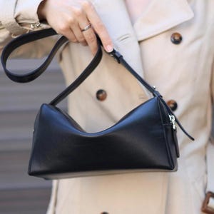 Pu&ograve; includere: Una borsa a tracolla rettangolare nera con finitura liscia, caratterizzata da un bordo superiore curvo, una chiusura a zip color argento e una tracolla nera regolabile fissata con minuteria in metallo argentato.