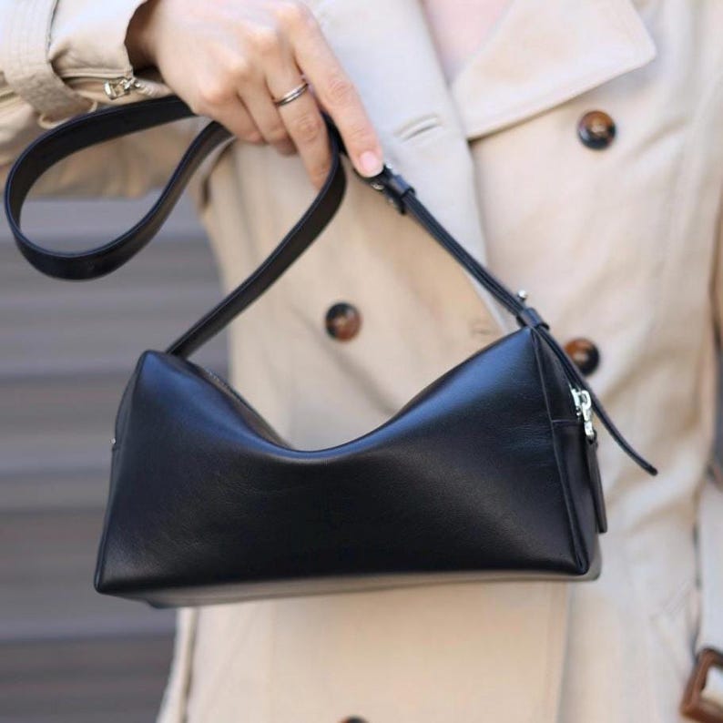 Pu&ograve; includere: Una borsa a tracolla rettangolare nera con finitura liscia, caratterizzata da un bordo superiore curvo, una chiusura a zip color argento e una tracolla nera regolabile fissata con minuteria in metallo argentato.