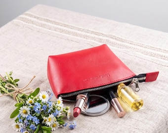 May include: A bright red leather cosmetic pouch with a black zip closure, featuring the embossed text LionLeather.com. Inside the pouch are a lipstick, a small round compact mirror, and a clear glass bottle filled with a golden liquid.