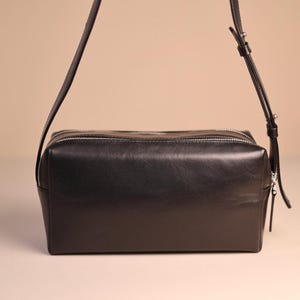Pu&ograve; includere: Una borsa a tracolla rettangolare in pelle nera con finitura liscia, dotata di due cerniere metalliche color argento sulla parte superiore e una tracolla regolabile con fibbia e dettagli in metallo color argento. Forma strutturata.