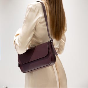 May include: A dark burgundy shoulder bag with a smooth, matte finish and a rectangular flap closure. The bag features a thin, adjustable strap with a silver-toned metal buckle, worn over the shoulder to rest against the side of the torso.