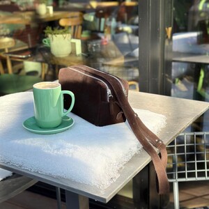 Pu&ograve; includere: Una borsa a tracolla rettangolare in camoscio marrone scuro con cerniere in metallo argentato e una tracolla regolabile abbinata, posizionata accanto a una tazza da caff&egrave; in ceramica verde menta chiaro su un piattino.