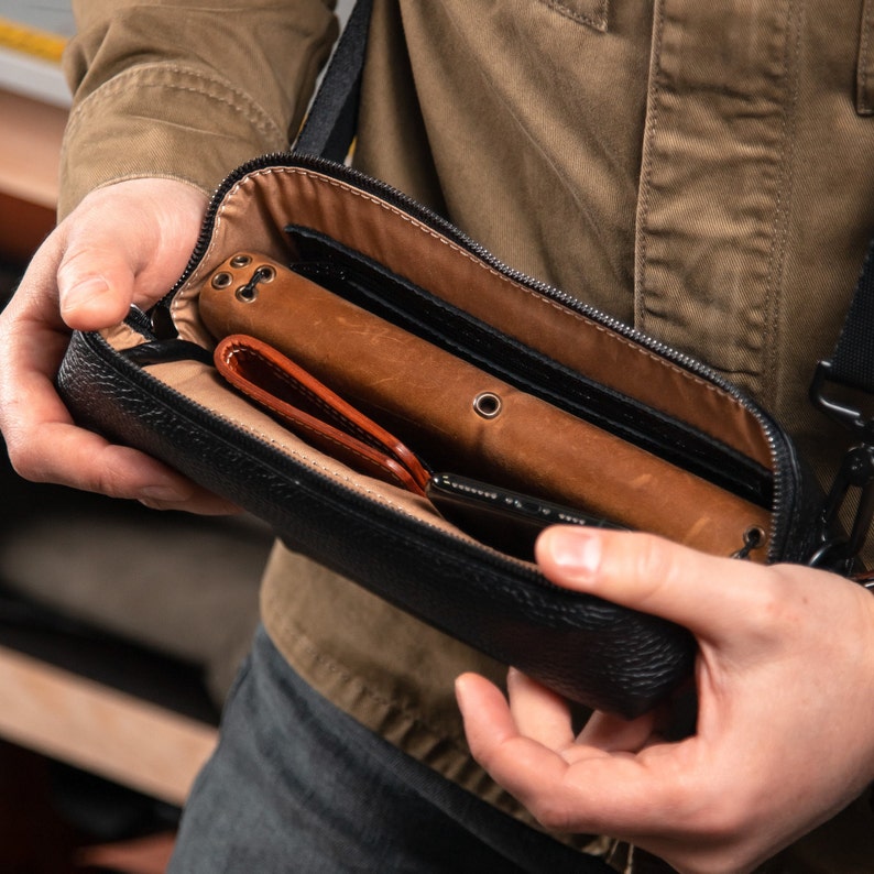 May include: A black textured leather crossbody bag with a zipper closure, containing a tan leather notebook cover with metal eyelets, a folded tan leather wallet, and a black smartphone inside the main compartment with a tan fabric lining.