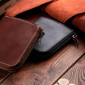 May include: Two small leather zipped pouches, one in a deep brown and one in black, featuring visible white stitching along the edges and silver-toned metal zips. The pouches have a rectangular shape with rounded corners and a smooth texture.