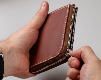 May include: A rectangular brown leather wallet featuring a silver-toned metal zip closure along the side. The wallet is constructed with visible white stitching along the edges and a brown fabric zip tape.