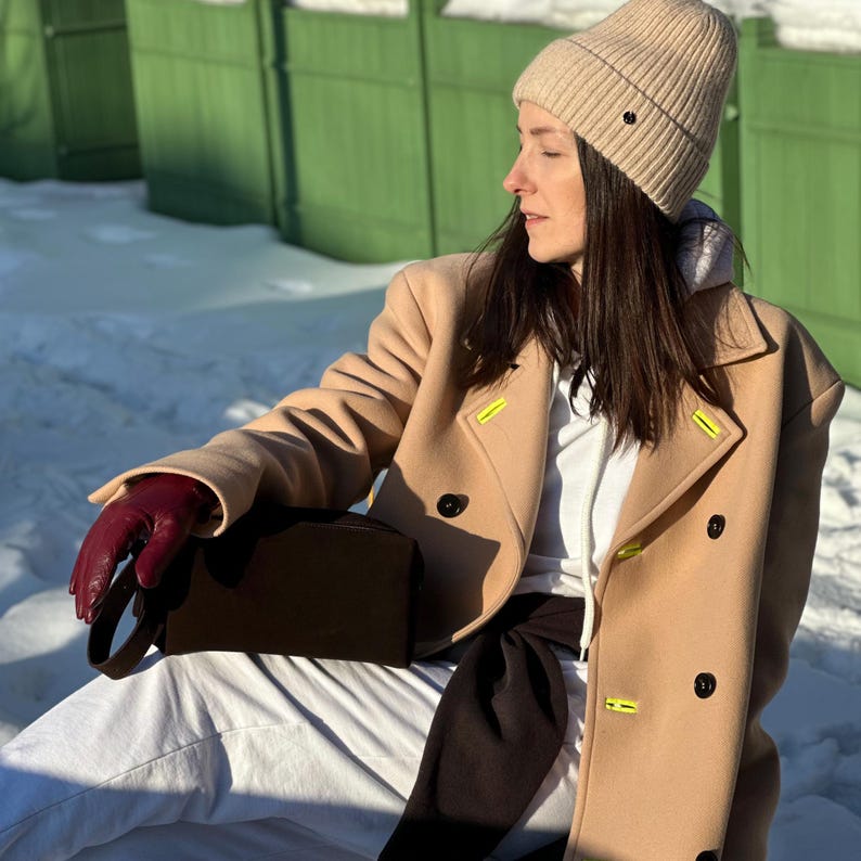Pu&ograve; includere: Un berretto in maglia a coste beige, un cappotto in lana doppiopetto color marrone chiaro con accenti di filo giallo neon sul bavero, una felpa con cappuccio bianca, un maglione nero legato in vita e un guanto in pelle rosso scuro con una pochette marrone.