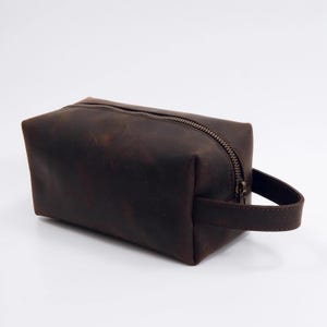 May include: A rectangular dark brown leather wash bag featuring a smooth matte finish, a single top zip closure with a metallic pull, and a matching leather side handle strap attached to the right side of the structured pouch.