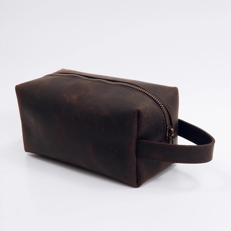May include: A rectangular dark brown leather wash bag featuring a smooth matte finish, a single top zip closure with a metallic pull, and a matching leather side handle strap attached to the right side of the structured pouch.