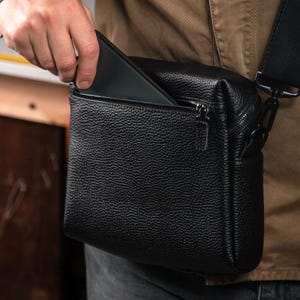 May include: A black crossbody bag with a textured, pebbled leather-like finish. The bag features a rectangular shape, a top zipper closure, and a front zippered pocket. A black adjustable strap is attached to the side with a metal clip hardware.