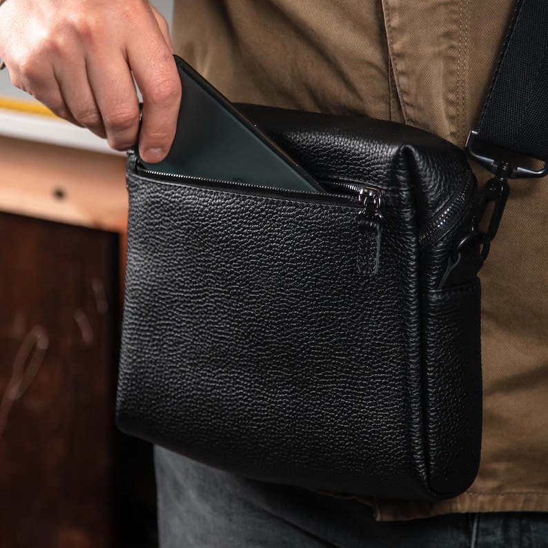 May include: A black crossbody bag with a textured, pebbled leather-like finish. The bag features a rectangular shape, a top zipper closure, and a front zippered pocket. A black adjustable strap is attached to the side with a metal clip hardware.