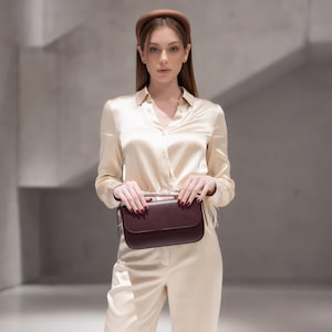 May include: A long-sleeved cream-colored satin button-down shirt and matching trousers, paired with a brown padded headband and a dark burgundy rectangular leather handbag with a flap closure and a top handle held in front of the body.