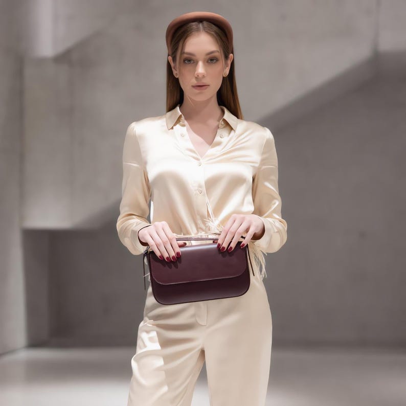 May include: A long-sleeved cream-colored satin button-down shirt and matching trousers, paired with a brown padded headband and a dark burgundy rectangular leather handbag with a flap closure and a top handle held in front of the body.