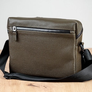 May include: An olive green crossbody bag with a textured, pebbled surface. The bag features a horizontal zippered pocket on the front with a silver-toned metal zipper pull and a wide, adjustable black fabric shoulder strap attached with black hardware.
