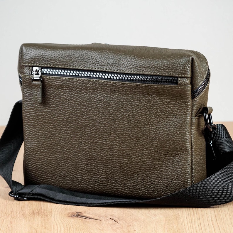 May include: An olive green crossbody bag with a textured, pebbled surface. The bag features a horizontal zippered pocket on the front with a silver-toned metal zipper pull and a wide, adjustable black fabric shoulder strap attached with black hardware.