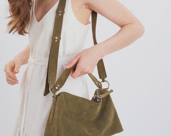 May include: An olive green suede shoulder bag featuring a rectangular silhouette, a top zipper closure with a matching pull tab, and an adjustable strap attached with silver-toned metal rings and rivets. The bag has a soft, matte texture.