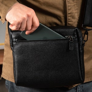 May include: A black textured leather crossbody bag featuring a zippered top compartment. The bag has a rectangular shape with a visible zipper pull and a black adjustable shoulder strap attached with metal hardware, shown with a smartphone.