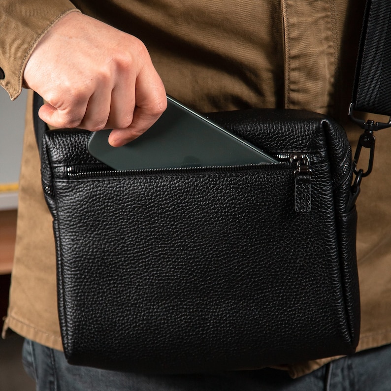 May include: A black textured leather crossbody bag featuring a zippered top compartment. The bag has a rectangular shape with a visible zipper pull and a black adjustable shoulder strap attached with metal hardware, shown with a smartphone.
