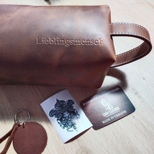 May include: A brown leather toiletry bag with a side handle and the embossed word "Lieblingsmensch". Included are a round leather keychain stamped "Lion Leather co.", a sticker with a lion illustration, and a card labelled "LION LEATHER".