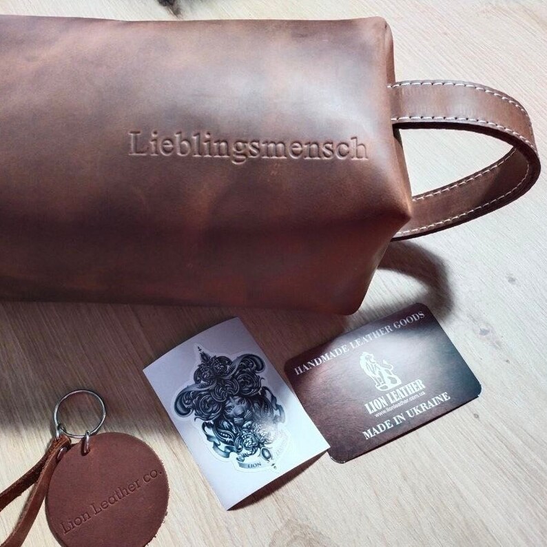 May include: A brown leather toiletry bag with a side handle and the embossed word "Lieblingsmensch". Included are a round leather keychain stamped "Lion Leather co.", a sticker with a lion illustration, and a card labelled "LION LEATHER".