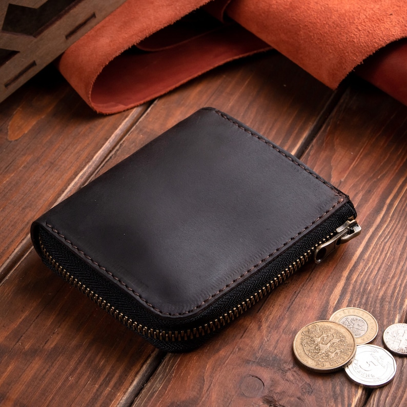 May include: A dark brown leather wallet with a smooth finish and a wrap-around metal zip closure. The wallet features visible stitching along the edges and a metallic zip pull. Several silver and gold-coloured coins are positioned nearby.