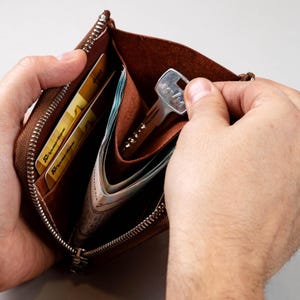 May include: A brown leather zipped wallet containing two gold-coloured credit cards, folded paper currency, and a silver-toned metal key. The wallet features a silver metal zip closure and an open interior compartment for organising small items.