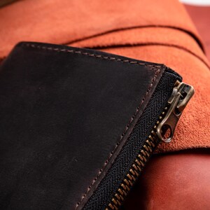 May include: A dark brown leather wallet featuring a matte finish and visible stitching along the edges. The wallet includes a black fabric zip tape with a metallic brass-toned zip pull, showcasing a textured surface and durable construction.