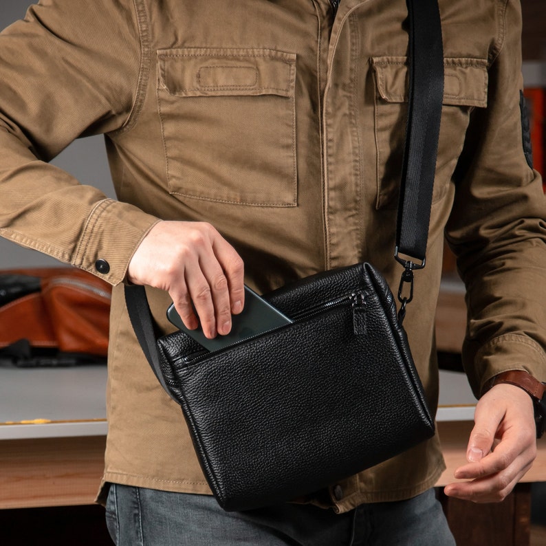 May include: A black textured leather crossbody bag featuring a zippered top closure and a wide black adjustable shoulder strap. The bag has a rectangular shape with a pebbled surface texture and a slim profile, designed for carrying small personal items.