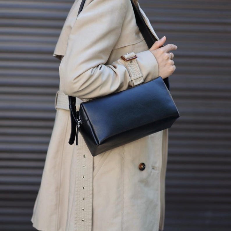Pu&ograve; includere: Una borsa a tracolla nera di forma rettangolare con finitura liscia, dotata di chiusura con zip color argento e tracolla nera regolabile abbinata, indossata sopra un trench beige con dettaglio del polsino abbottonato.