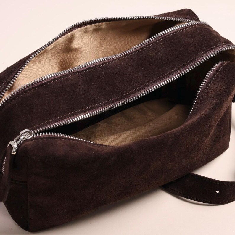 May include: A dark brown suede rectangular bag with a silver metal zip closure. The interior is lined with a light beige fabric. The bag features a structured shape with a single strap visible at the bottom right corner of the frame.