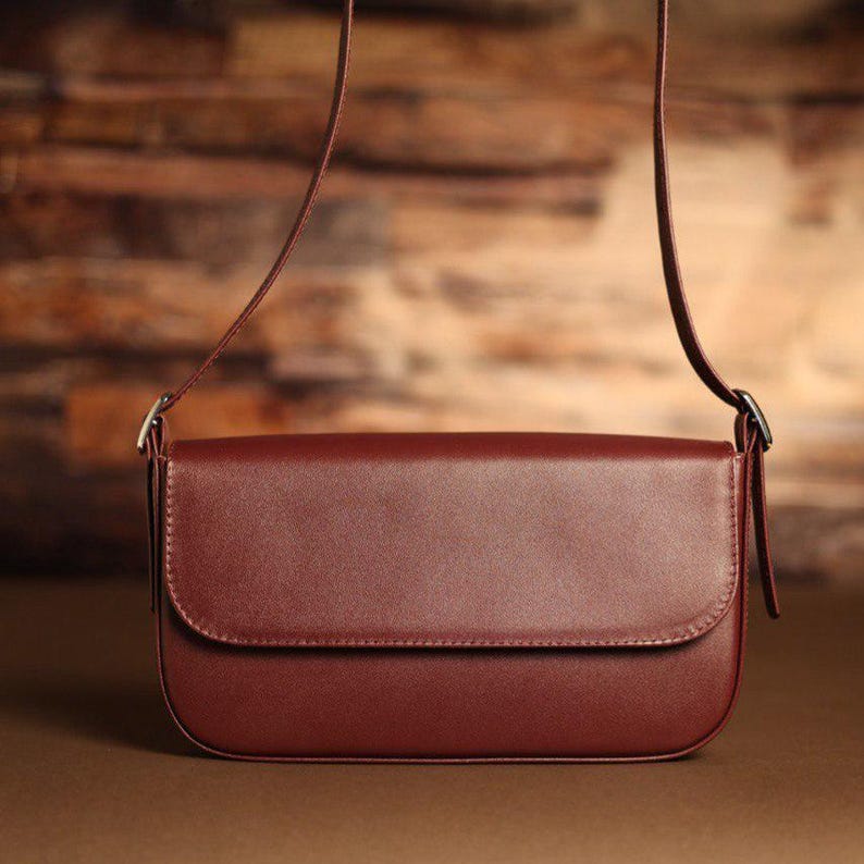 May include: A deep burgundy rectangular shoulder bag with a smooth, solid-colored exterior, a rounded bottom edge, a fold-over flap closure, and a thin, matching adjustable shoulder strap attached with silver-toned metal hardware on both sides.